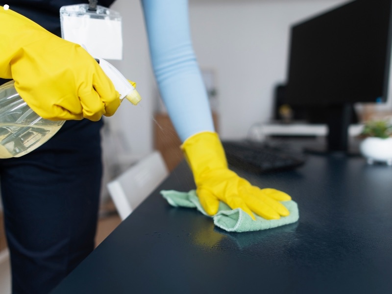 Cleaner disinfecting an office desk beside a monitor